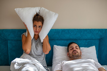 Young couple have problem with man's snoring. Heterosexual couple in bed, man sleeps and snoring with mouth open, while a tired woman irritated by snoring sitting on bed with a pillow on her head.