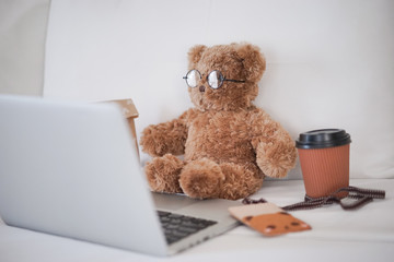 Teddy bear working concept. A cute teddy bear working in bed with a grey laptop and a cup of hot coffee