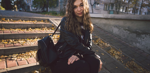 A young beautiful woman in casual clothes is sitting on the steps with leaves in the urban district.