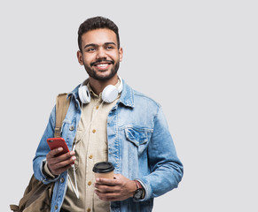 Young handsome man with backpack holding smart phone and coffee. Smiling student going on a travel. Isolated on gray background - obrazy, fototapety, plakaty