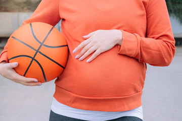 Pregnant girl in orange sweatshirt with a basketball ball in hand. Pregnant outdoor training.
