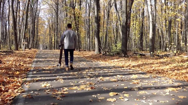 the guy climbs up the hill on the asphalt covered with leaves, with a longboard in his hands