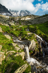 A glacial river running out of the high alpine mountains through an alpine meadow  - 297072990