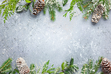 Christmas or winter background with a border of green and frosted evergreen branches and pine cones on a grey vintage board. Flat lay
