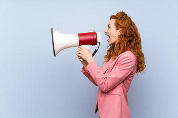 Redhead woman in suit over isolated blue wall shouting through a megaphone