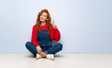 Redhead woman with overalls sitting on the floor showing and lifting a finger in sign of the best