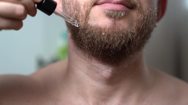 Close-up shot of handsome bearded man staying in the bathroom in front of mirror and putting droplets of fresh aromatic essential oil on his dry beard. Relaxing procedures after beard shaving with