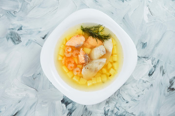 Homemade transparent fish soup with sea bass, salmon and shrimp in a white bowl on a gray background. Seafood