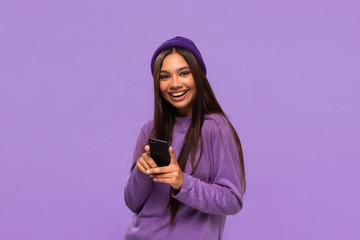 Smiling african-american girl in a hat and sweater holding smartphone isolated over purple background.