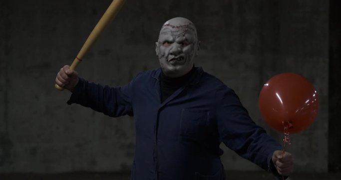 Extremely dangerous man in mask of movie maniac attacks spectator with baseball bat, psycho in parking lot, Halloween holiday, creepy costume, October walk, red ball in his hand, alien from hell
