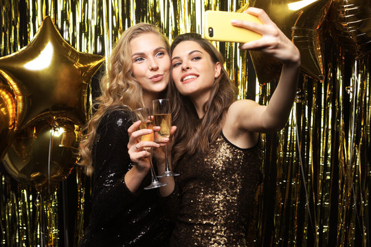 Image of two attractive girls in stylish outfit holding smartphone and taking selfie photo on gold background.