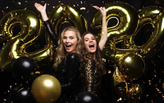 Happy gorgeous girls in stylish sexy party dresses holding gold 2020 balloons, having fun at New Year's Eve Party.