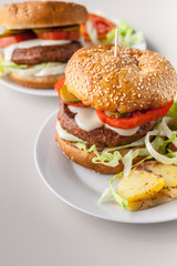 Two homemade hamburgers with a juicy meat cutlet, parmesan cheese, green salad, tomatoes and fried potatoes on a light background. Vertical shot