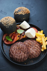 Grilled beef burger cutlets with french fries, tomatoes, cheese and black buns on a metal tray, closeup on a black stone surface