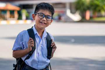 A cute Asian elementary school boy wearing glasses and a white school uniform, carrying a black backpack, standing in front of the school.