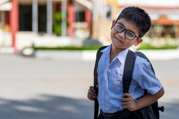 A cute Asian elementary school boy wearing glasses and a white school uniform, carrying a black backpack, standing in front of the school.