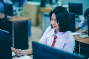 Fototapeta na wymiar Smiling Asian female high school student in white uniform is enjoying social media in computer room.