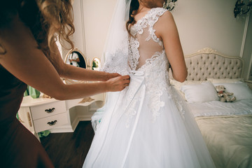 Gorgeous bride in white luxury dress is getting ready for wedding.  Woman putting on dress. Stylish bride getting dressed in modern gown with bridesmaid. Morning preparation before wedding ceremony.