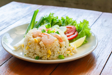 Close up fried rice with shrimp, egg and green onion on white plate serve with blurry cucumber, red tomato, lettuce in cafe and restaurant.