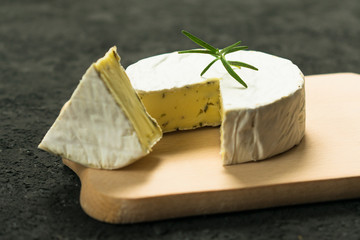 Camembert cheese with greens decorated with a sprig of rosemary on a wooden board on a black background.