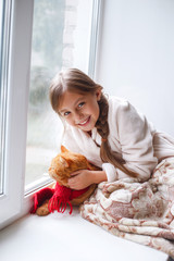 Little girl in teddy robe sitting on a window sill with pet. Lazy weekend with red cat at home.