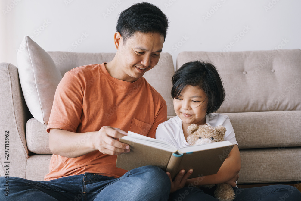 Wall mural Asian father and daughter reading book while sitting in the living room at her home.