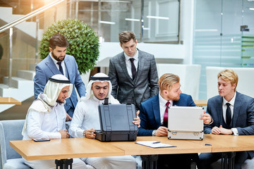 Fototapeta na wymiar Caucasians in suits evaluate money by sheikhs, sheikhs in white suits look at modern technology, opening black case. Business concept
