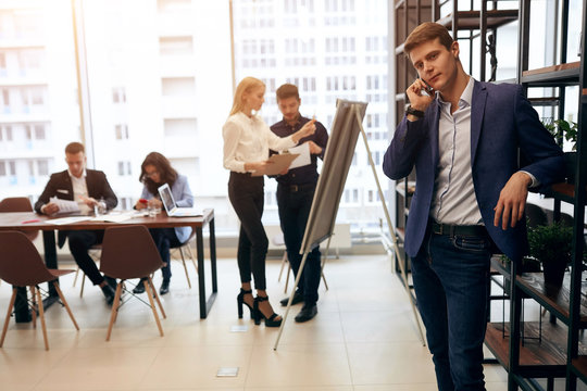 young attractive elegant man in blue suit consulting clients by smartphone while his colleagues working on project. working atmosphere
