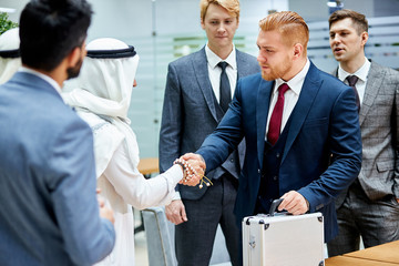 Fototapeta premium Sheikhs making deal with caucasian man in suit. Two businessmen shaking hands to each other. Business center background