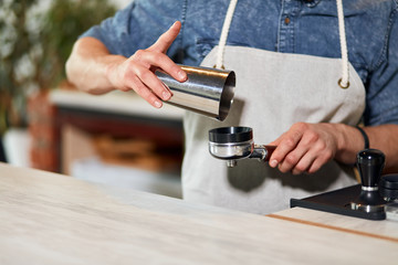 Hands of male barista holds metal coffee pot and tamper over wooden light coloured counter