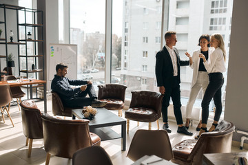 Business people meeting, talking while standing in front of the panorama window, while handsome cheerful man sitting on the armchair and using his tablet. copy space.conversation concept - 297119973