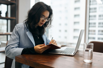 attractive cute office worker writing something on her notepad while sitting at the table in the office with panorama window. close up side view photo. copy space