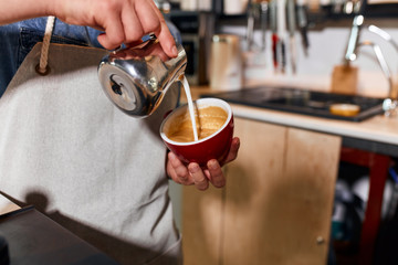 Hands of skillful young bartender pouring steamed milk from metal jug, making hot latte, holding red big cup, coffee making concept