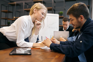 blonde attractive girl with long straight hair helping her colleague to make some notes during the meeting, close up side view photo. girl consulting his new employee, giving recommendatons