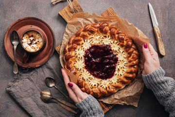 Cottage cheese pie with strawberry jam in female hands, a cup of coffee or hot chocolate with marshmallows on a dark rustic background. Top view, flat lay.