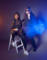 Fototapeta premium Black and white concept. Dark-skinned girl in black clothes sitting on a white stool. A man with white skin smokes VAPE. Communication of different people. African girl with a white phone.