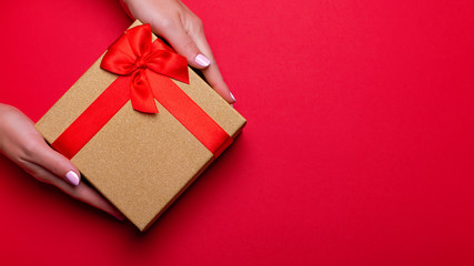 Woman manicured hands holding red and golden wrapped present or giftbox on deep red background, copy space, top view, flat lay. Background for Valentine's Day, Mother's Day.