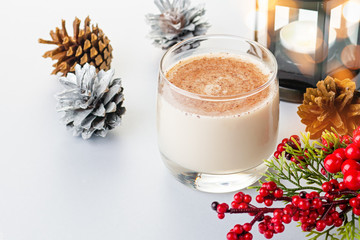 Traditional Christmas cocktail Eggnog with eggs, alcohol, grated nutmeg and cinnamon closeup. Sweet traditional drink on grey table with beige decorations, burning lanterns and pine cones