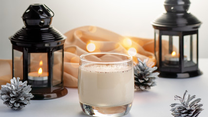 Traditional Christmas cocktail Eggnog with eggs, alcohol, grated nutmeg and cinnamon closeup. Sweet traditional drink on grey table with beige decorations, burning lanterns and pine cones