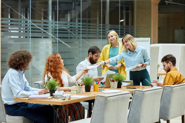 Cheerful girl with long blonde hair dressed in light blue shirt and casual jeans, holds documents, smiles happily, gives white shit of paper to red haired colleague in design studio, horizontal shot