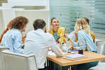 Hard working team of bright designers brainstorming in modern office, pretty blonde woman sits at table, dressed in colourful clothres, holds pencil, keeps hand on gentle cheek, looks away