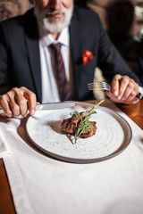 Chief cook served tasty dish on table. hardened male hands hold silver knife and fork ready to cut a piece of fried meat, specially cooked meat on white round plate. Down shot, close up