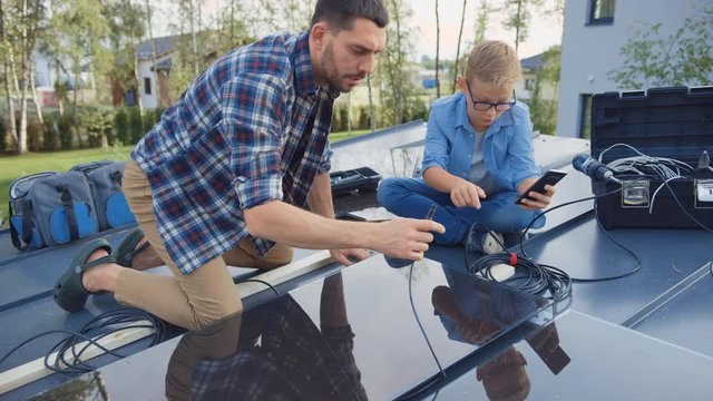Father and Son Installing Solar Panels to a Metal Basis. They Work with Wiring on a House Roof on a Sunny Day. Boy is Looking at a Manual on a Smartphone. Father Connects the Plugs.