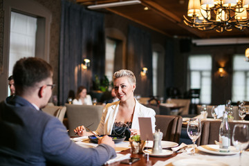 Close up of glorious optimistic young lady wears white unbuttoned jacket and black blouse, admiringly looks at business partner with eyes full of happiness at restaurant table