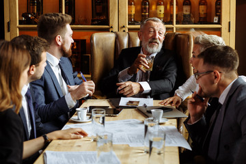 Businessmen have casual conversation in a cozy office. Cheerful gray-haired man sits in a large leather chair at the head of woden table and holds a glass of fine whiskey