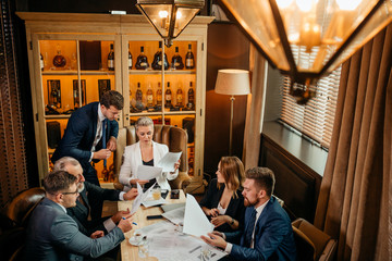 doubtful young man in formal suit attentively examining papers sitting at light coloured table in small study, background of black out brown curtains, top view