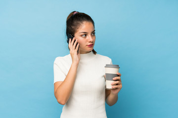 Young brunette girl over isolated blue background holding coffee to take away and a mobile