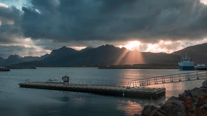  pier on the background of dramatic landscape at sunset. The rays of the sun make their way through the clouds , boats and mountain peaks. Lofoten Islands . Norway. 