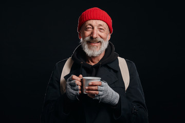 Bearded beggar male smiling, happy even living in street. Cheerful homeless man with mug of hot tea isolated over black background