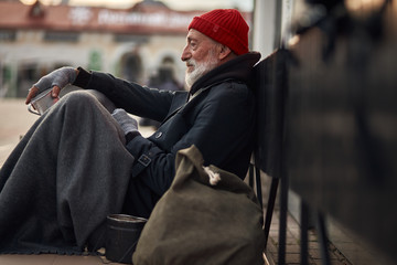 Side view on beggar male on street begging, waiting help by citizens. Senior homeless man in old...